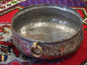 Antique hand-hammered copper bowl with a brass ring handle, resting on a traditional Turkish rug. Shows the interior depth and textured exterior patina
